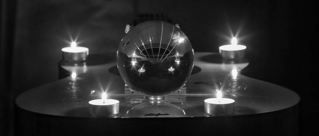 Ein Schwarz-Weiß-Foto einer Glaskugel auf einem Tisch mit brennenden Kerzen darum herum.