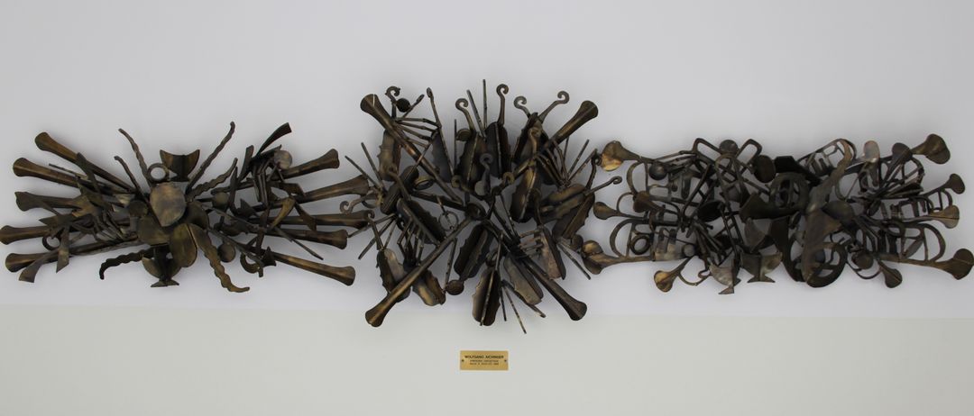 Eine Bronzeskulptur mit einer abstrakten Anordnung verschiedener Musikinstrumente, einschließlich Violinen, Violoncelli und Trompeten, ist auf einem weißen Hintergrund zu sehen. Unten befindet sich ein Etikett mit der Aufschrift 'WOLFGANG AUCHINGER' und zusätzlichem Text in kleinerer Schrift.