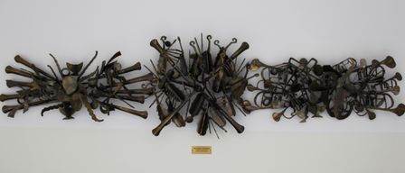 Eine Bronzeskulptur mit einer abstrakten Anordnung verschiedener Musikinstrumente, einschließlich Violinen, Violoncelli und Trompeten, ist auf einem weißen Hintergrund zu sehen. Unten befindet sich ein Etikett mit der Aufschrift 'WOLFGANG AUCHINGER' und zusätzlichem Text in kleinerer Schrift.