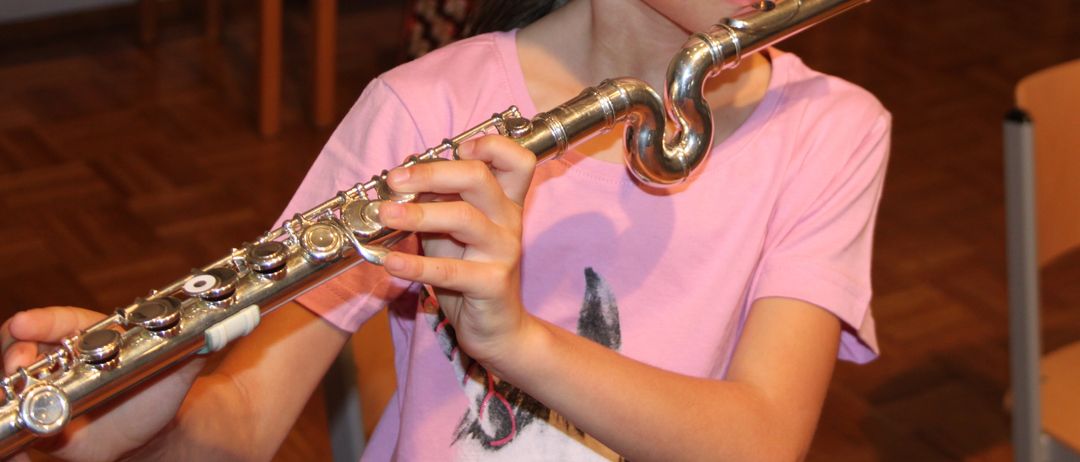 Ein junges Mädchen spielt Flöte, sitzt in einem Raum mit Holzstühlen im Hintergrund.