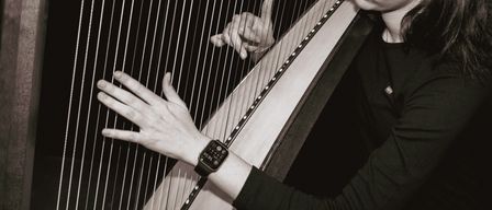 Eine Frau in Schwarz spielt eine Harfe, ihre Hände auf den Saiten positioniert, eine Hand hält den Hals des Instruments.