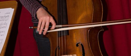 Ein junges Mädchen mit geflochtenen Haaren hält ein Cello. Sie steht vor einem roten Vorhang, wobei ein Notenständer links sichtbar ist.