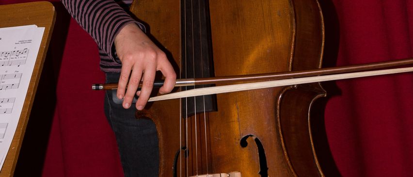 Ein junges Mädchen mit geflochtenen Haaren hält ein Cello. Sie steht vor einem roten Vorhang, wobei ein Notenständer links sichtbar ist.