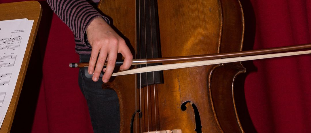 Ein junges Mädchen mit geflochtenen Haaren hält ein Cello. Sie steht vor einem roten Vorhang, wobei ein Notenständer links sichtbar ist.