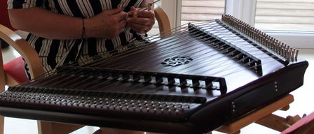 Eine Frau mit Brille und Halskette spielt eine Zither auf einem Balkon mit Holzdeck und Tisch.