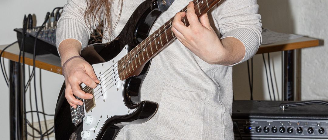 Eine Frau in einem grauen Oberteil spielt eine schwarze Gitarre mit einem roten Gurt, neben einem schwarzen Verstärker in einem Studio stehend.