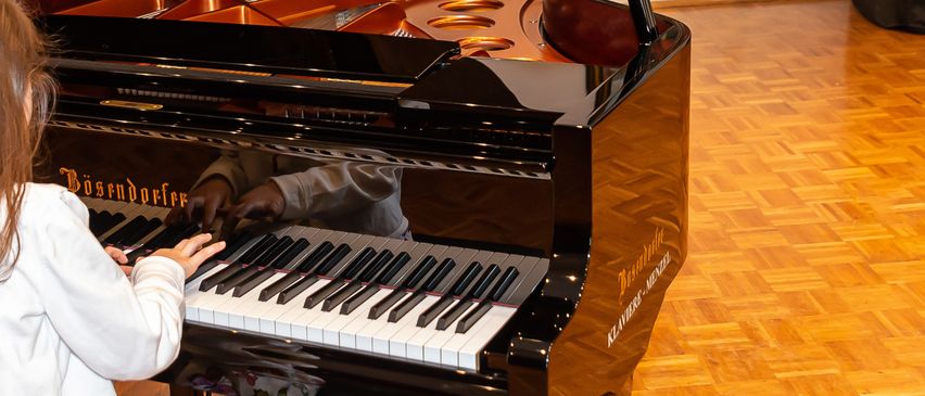 Ein kleines Mädchen sitzt auf einem Hocker vor einem Flügel und spielt mit ihren Fingern. Dahinter befindet sich eine weiße Wand mit einer geschlossenen Tür und einem gerahmten Gemälde. In der Nähe steht eine schwarze Klavierabdeckung.