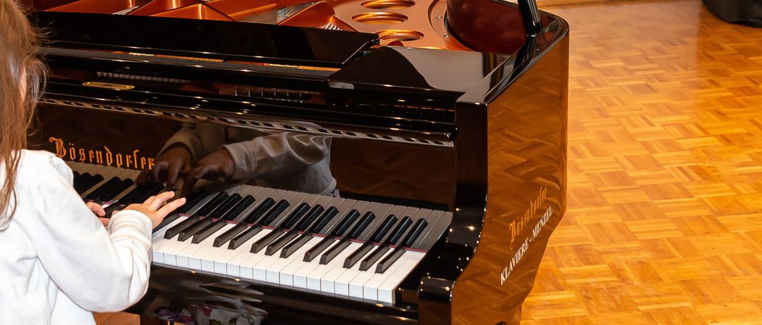 Ein kleines Mädchen sitzt auf einem Hocker vor einem Flügel und spielt mit ihren Fingern. Dahinter befindet sich eine weiße Wand mit einer geschlossenen Tür und einem gerahmten Gemälde. In der Nähe steht eine schwarze Klavierabdeckung.