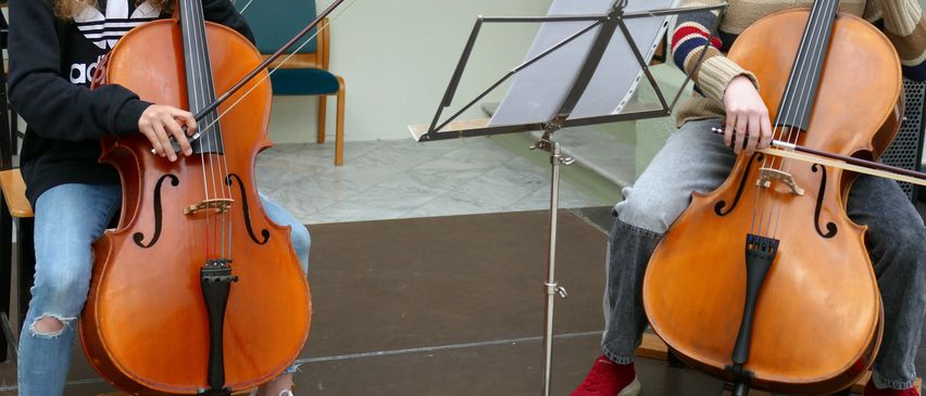 Zwei Frauen spielen Cello, sitzend in einem Raum. Sie haben Notenständer vor sich. Dahinter steht ein Stuhl und eine Wand mit einem Bilderrahmen.