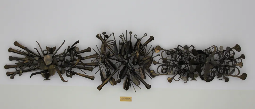 Eine Bronzeskulptur verschiedener Musikinstrumente ist an einer weißen Wand montiert. Darunter befindet sich eine kleine Plakette mit dem Text 'WOLFGANG WOLFF'. Vor der Wand stehen schwarze Stühle.