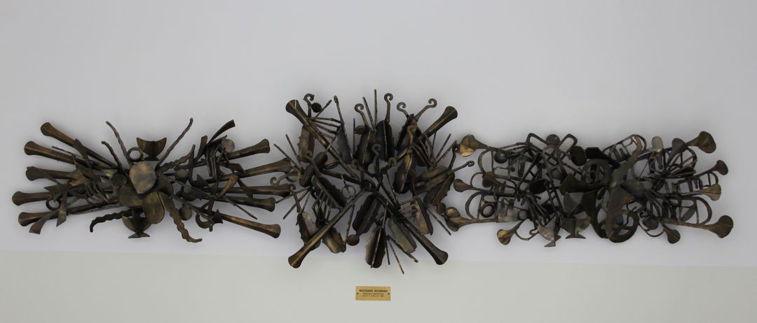 Eine Bronzeskulptur verschiedener Musikinstrumente ist an einer weißen Wand montiert. Darunter befindet sich eine kleine Plakette mit dem Text 'WOLFGANG WOLFF'. Vor der Wand stehen schwarze Stühle.