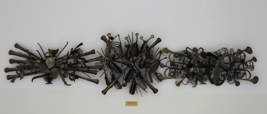 Eine Bronzeskulptur verschiedener Musikinstrumente ist an einer weißen Wand montiert. Darunter befindet sich eine kleine Plakette mit dem Text 'WOLFGANG WOLFF'. Vor der Wand stehen schwarze Stühle.