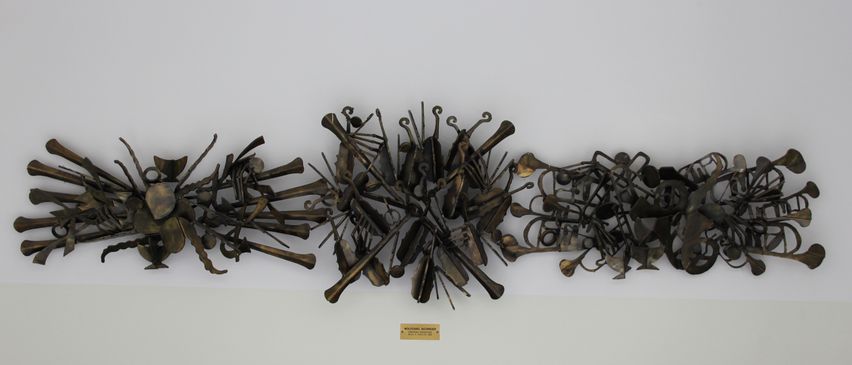Eine Bronzeskulptur verschiedener Musikinstrumente ist an einer weißen Wand montiert. Darunter befindet sich eine kleine Plakette mit dem Text 'WOLFGANG WOLFF'. Vor der Wand stehen schwarze Stühle.