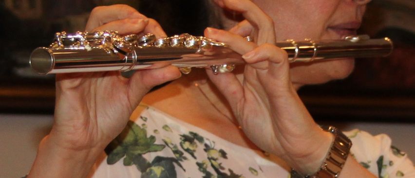 Eine Frau in einem weißen Blumenshirt spielt Flöte vor einem Notenständer. Dahinter spielt eine Person Akkordeon.