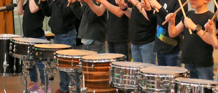 Eine Gruppe junger Jungen in schwarzen Shirts und Jeans spielt in einem Raum Trommeln. Sie halten Drumsticks und lächeln. Dahinter ist ein verschwommenes Bild einer Person, die ein Foto macht.
