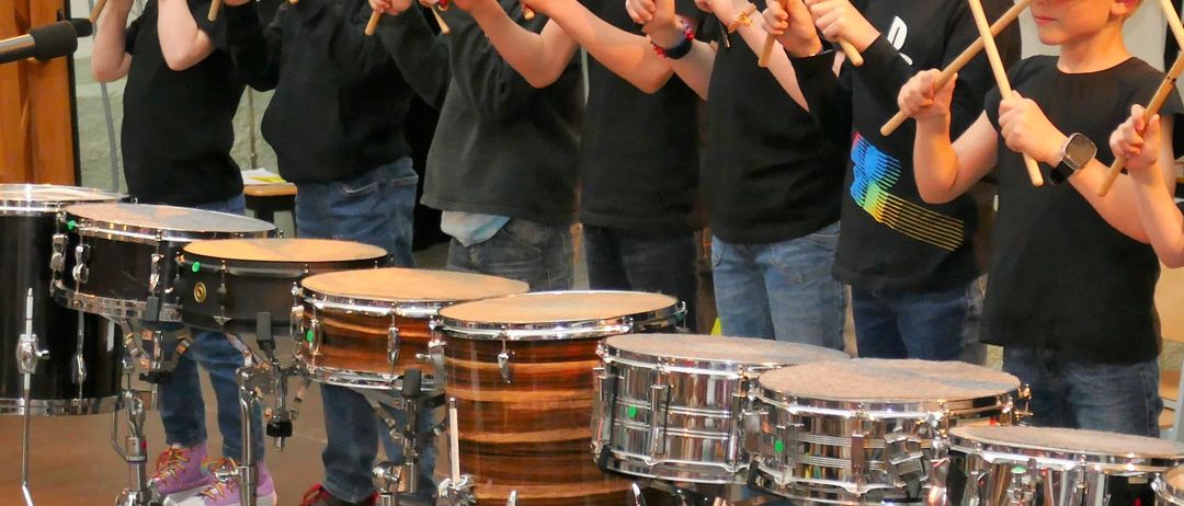 Eine Gruppe junger Jungen in schwarzen Shirts und Jeans spielt in einem Raum Trommeln. Sie halten Drumsticks und lächeln. Dahinter ist ein verschwommenes Bild einer Person, die ein Foto macht.