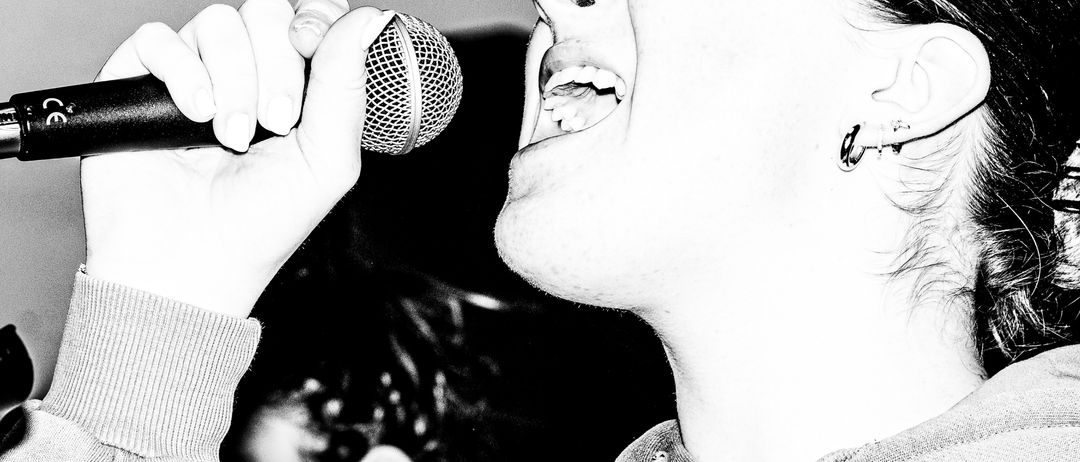 Eine Frau singt in ein Mikrofon, ihre Augen geschlossen, während eine weitere Person mit einem Pferdeschwanz im Hintergrund zu sehen ist.