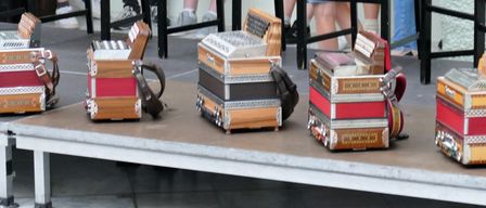 Mehrere Akkordeons in Rot, Schwarz und Braun stehen auf einer Bühne. Dahinter sitzen Menschen auf Stühlen und tragen Sneakers und Socken.