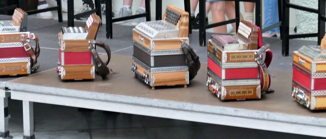 Mehrere Akkordeons in Rot, Schwarz und Braun stehen auf einer Bühne. Dahinter sitzen Menschen auf Stühlen und tragen Sneakers und Socken.
