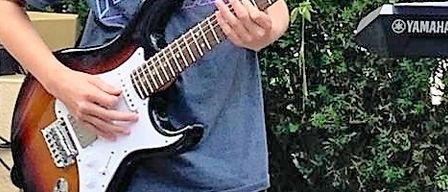 Zwei junge Musiker treten draußen auf. Ein Junge spielt E-Gitarre und ein Mädchen spielt Keyboard. Sie stehen vor Büschen mit Bäumen im Hintergrund.