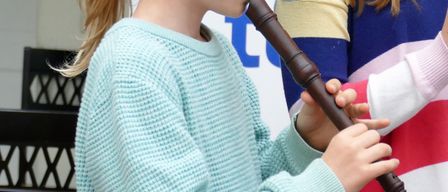 Zwei junge Mädchen spielen Blockflöten in einem Raum. Ein Mädchen trägt ein türkisfarbenes Sweatshirt, das andere ein mehrfarbiges Oberteil. Sie stehen neben einem Notenständer.