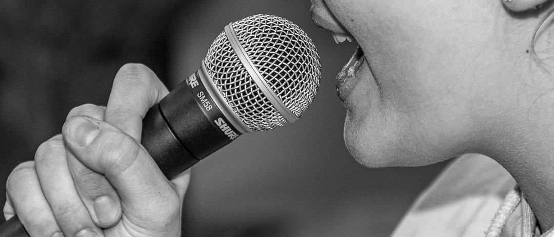 Ein Schwarz-Weiß-Foto einer Frau, die in ein Mikrofon singt. Sie hat ihren Mund geöffnet und ihre Augen geschlossen, mit einem ernsten Ausdruck.