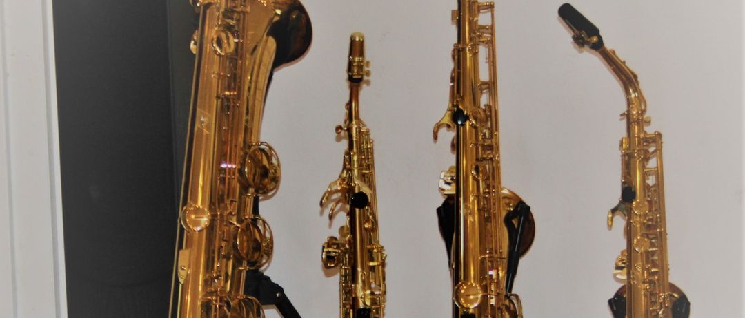 Vier goldene Saxophone sind auf Stativen vor einer weißen Wand ausgestellt. Hinter dem linken Saxophone steht ein schwarzer Lautsprecher.