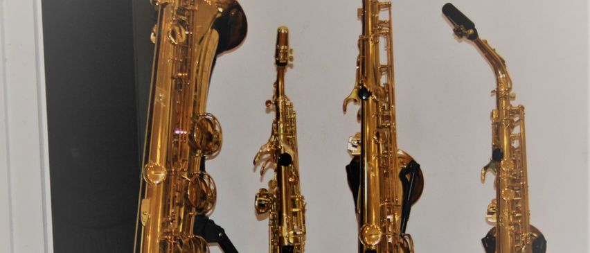 Vier goldene Saxophone sind auf Stativen vor einer weißen Wand ausgestellt. Hinter dem linken Saxophone steht ein schwarzer Lautsprecher.