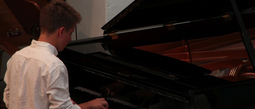 Ein junger Mann in einem weißen Hemd spielt mit seiner rechten Hand ein schwarzes Klavier. Das Klavier ist geöffnet, und das Innere ist sichtbar. Der Raum ist schwach beleuchtet.