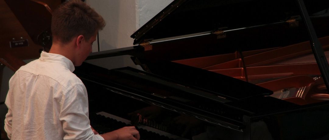 Ein junger Mann in einem weißen Hemd spielt mit seiner rechten Hand ein schwarzes Klavier. Das Klavier ist geöffnet, und das Innere ist sichtbar. Der Raum ist schwach beleuchtet.