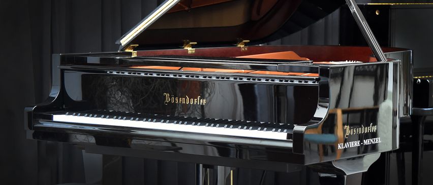 Ein schwarzer Flügel mit goldenen Akzenten und einem goldenen Pedal wird auf einer Bühne ausgestellt. Der Deckel des Klaviers ist geöffnet und zeigt das Innere und die Tasten. Ein weiteres Klavier ist im Hintergrund teilweise sichtbar.