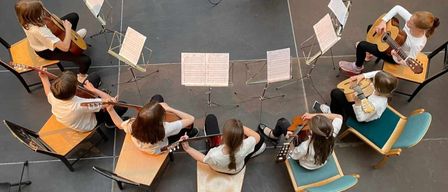 A group of young girls are playing guitars in a circle. They are sitting on benches, each holding a guitar. Music sheets are placed on stands in front of them. The room has a tiled floor and gray walls.