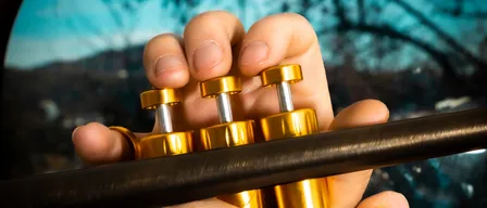 Eine Person spielt eine Trompete und hält sie mit beiden Händen. Die Trompete ist golden und hat eine glänzende Oberfläche. Die Person trägt einen Ring am Finger.