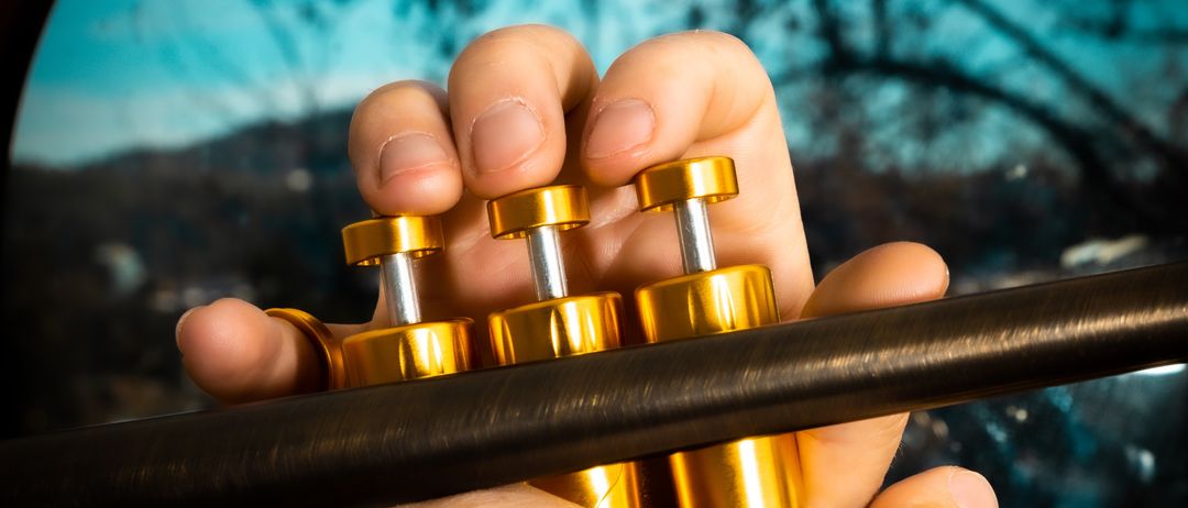 Eine Person spielt eine Trompete und hält sie mit beiden Händen. Die Trompete ist golden und hat eine glänzende Oberfläche. Die Person trägt einen Ring am Finger.