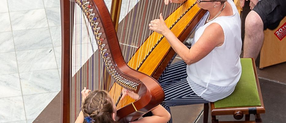 Eine Gruppe von Menschen sitzt auf Stühlen und beobachtet eine Frau, die Harfe spielt, während ein Mann neben ihr sitzt. Dahinter steht ein Klavier und ein Gitarrenkoffer.