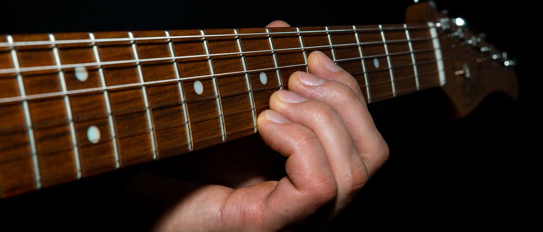 Ein Nahaufnahme des Halses einer Gitarre und einer Hand, die sie spielt. Der Hals der Gitarre hat eine braune Oberfläche mit weißen Punkten und Metallbünden. Die Hand ist positioniert, um einen Akkord zu spielen.