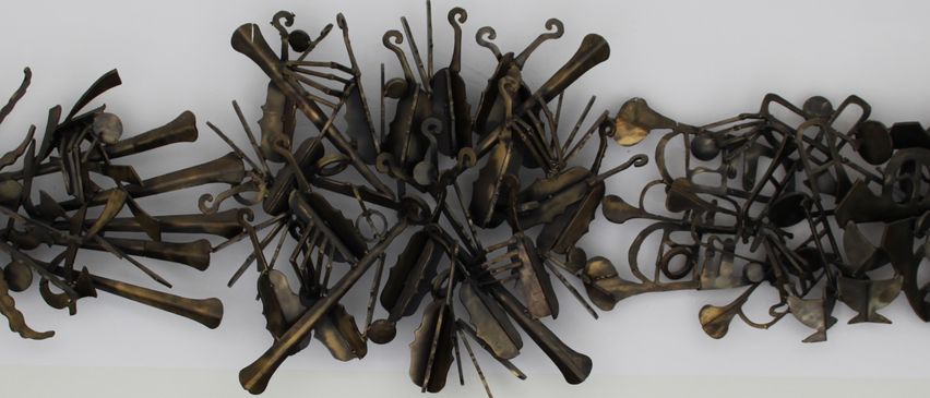 Eine Bronzeskulptur aus Musikinstrumenten, die in einem kreisförmigen Muster angeordnet sind. Verschiedene Instrumente wie Violinen, Trompeten und Celli sind kunstvoll verbunden. Eine Plakette am unteren Rand zeigt, dass sie von Wolfgang A. Hochinger stammt.