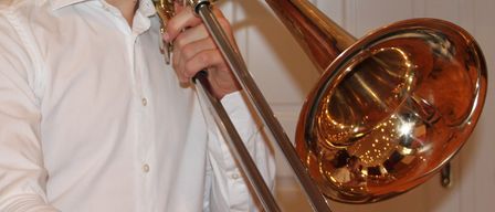 Ein junger Mann mit Brille hält eine Posaune, lächelt und steht in einem Raum mit weißer Tür und Bilderrahmen.