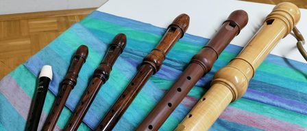 Five wooden flutes of varying sizes are arranged on a multicolored cloth, with a white object to the left. The flutes have brown and beige finishes, and the background is white.