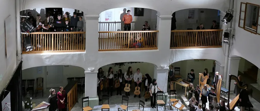 Eine Gruppe von Menschen in einem mehrstöckigen Raum, einige stehen auf Balkonen, andere spielen Instrumente und singen. Der Raum hat weiße Wände und Holzbalkone.