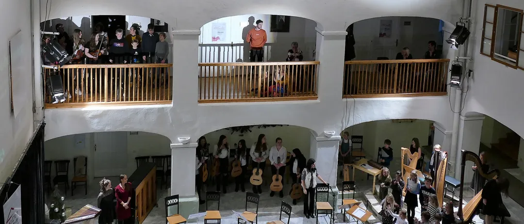 Eine Gruppe von Menschen in einem mehrstöckigen Raum, einige stehen auf Balkonen, andere spielen Instrumente und singen. Der Raum hat weiße Wände und Holzbalkone.