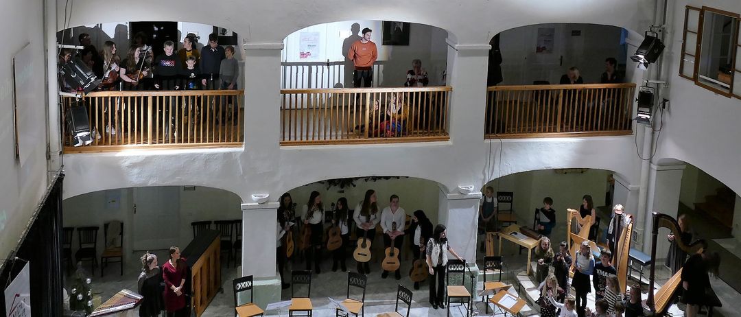 Eine Gruppe von Menschen in einem mehrstöckigen Raum, einige stehen auf Balkonen, andere spielen Instrumente und singen. Der Raum hat weiße Wände und Holzbalkone.