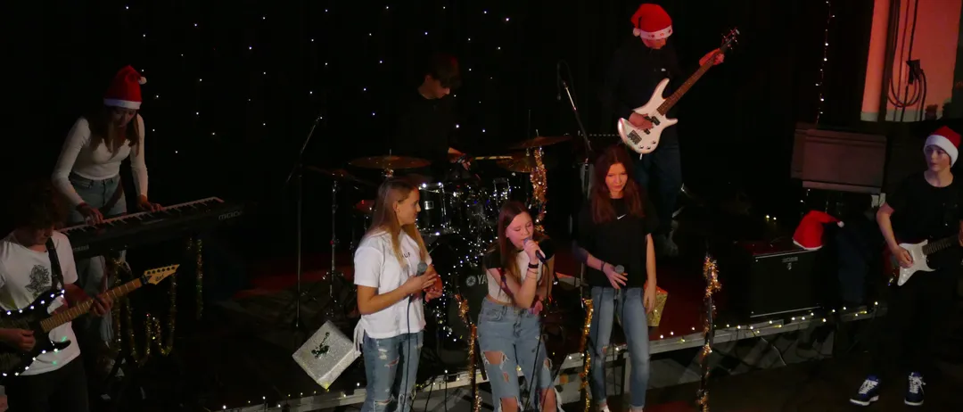 Drei junge Frauen singen auf der Bühne mit einem Gitarristen im Hintergrund. Sie stehen vor einem Schlagzeug und einem Mikrofon. Die Bühne ist mit Weihnachtslichtern dekoriert.