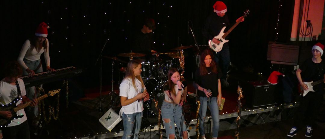 Drei junge Frauen singen auf der Bühne mit einem Gitarristen im Hintergrund. Sie stehen vor einem Schlagzeug und einem Mikrofon. Die Bühne ist mit Weihnachtslichtern dekoriert.