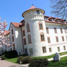Ein großes weißes Gebäude mit rotem Dach und Turm steht auf einem grünen Rasen, umgeben von Bäumen und Büschen.