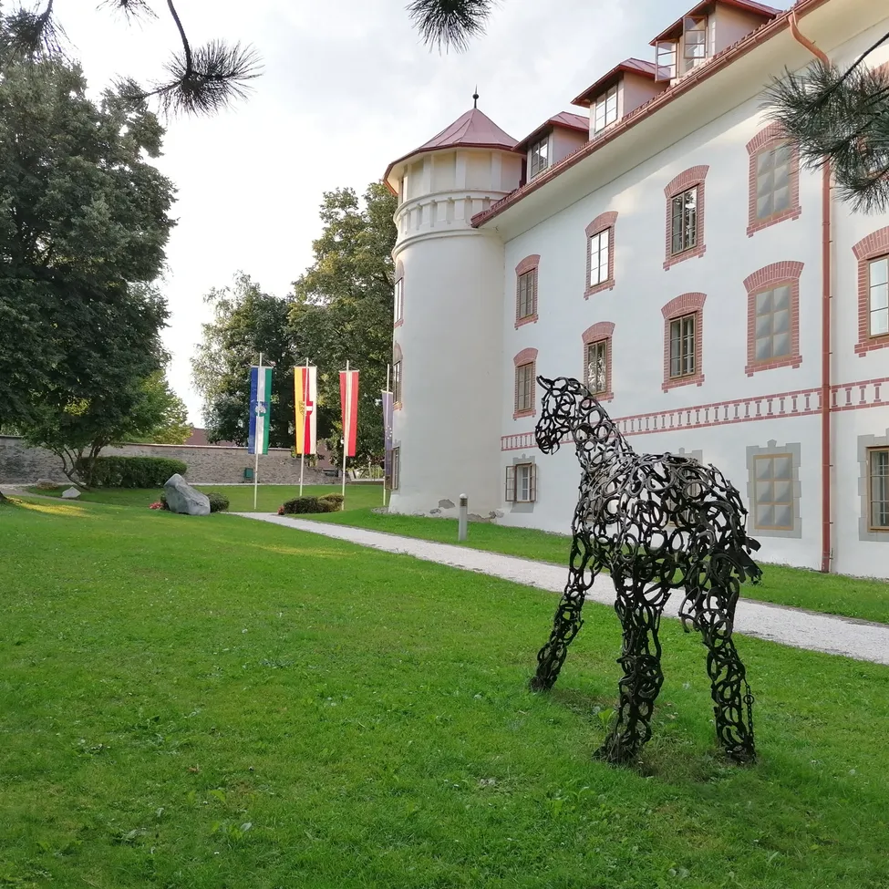 Eine Giraffenskulptur steht vor einem großen weißen Gebäude mit vielen Fenstern. Hinter der Skulptur befinden sich drei Fahnen. Das Gelände ist mit Gras bedeckt.