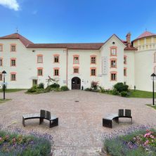 Ein großes weißes Gebäude mit rotem Dach und orangefarbenen Fenstereinfassungen. Zwei Bänke stehen davor.