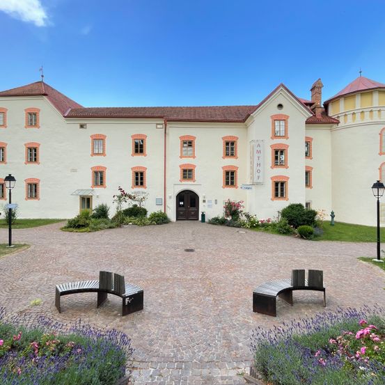 Ein großes weißes Gebäude mit rotem Dach und orangefarbenen Fenstereinfassungen. Zwei Bänke stehen davor.