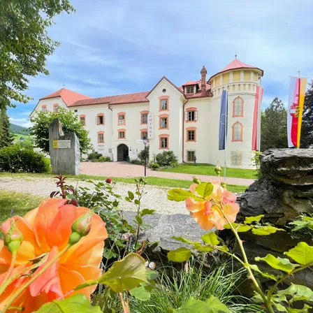 Ein weißes Gebäude mit roten Dächern steht vor einem Garten mit orangefarbenen Blumen und Fahnen an Stangen. Bäume und Büsche sind im Hintergrund.