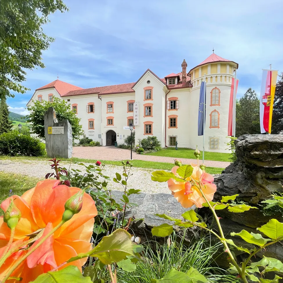 Ein weißes Gebäude mit roten Dächern steht vor einem Garten mit orangefarbenen Blumen und Fahnen an Stangen. Bäume und Büsche sind im Hintergrund.